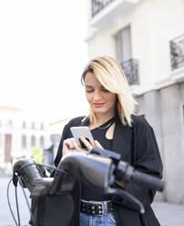 Blonde Frau benutzt Mobiltelefon vor einem Elektrofahrzeug - JCCMF02211
