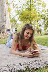 Lächelnde Frau mit Mobiltelefon auf einer Decke im Park - JRVF00560