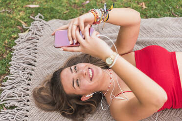 Happy woman listening music through headphones while using smart phone on blanket in park - JRVF00562