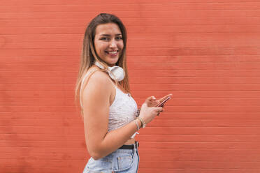 Beautiful woman with mobile phone standing by orange wall - JRVF00569