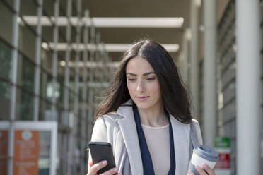 Geschäftsfrau, die einen wiederverwendbaren Kaffeebecher hält und ein Mobiltelefon benutzt - EIF01017