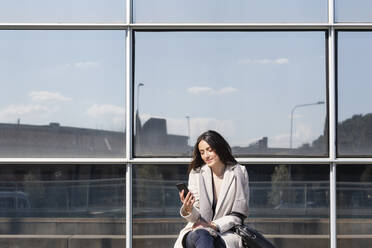 Geschäftsfrau, die ein Mobiltelefon benutzt, während sie vor einem Gebäude sitzt - EIF01062
