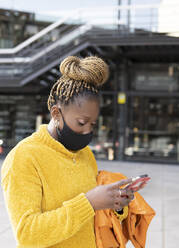 African woman wearing protective face mask using smart phone in city - JCCMF02225