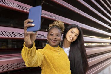 African woman taking selfie with female friend through smart phone by wall - JCCMF02230