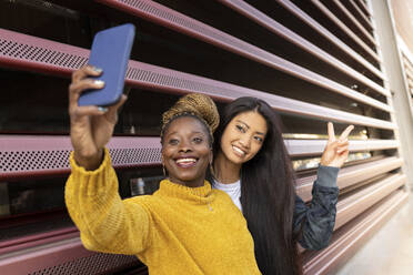 Young woman taking selfie with female friend showing peace sign by wall - JCCMF02231