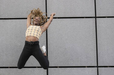 African woman jumping with arms raised by wall - JCCMF02247