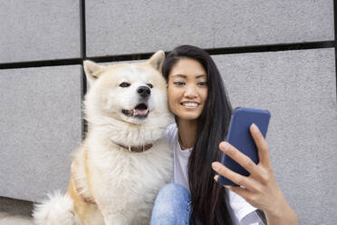 Hipster-Frau macht Selfie mit Akita-Hund durch Handy an der Wand - JCCMF02263