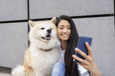 Hipster woman taking selfie with Akita dog through mobile phone by wall - JCCMF02263