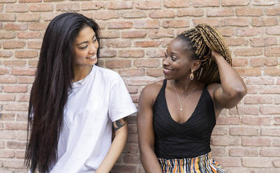 African woman with hand in hair looking at female friend by wall - JCCMF02280