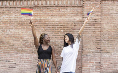 Lesbian couple waving rainbow flag by wall - JCCMF02282