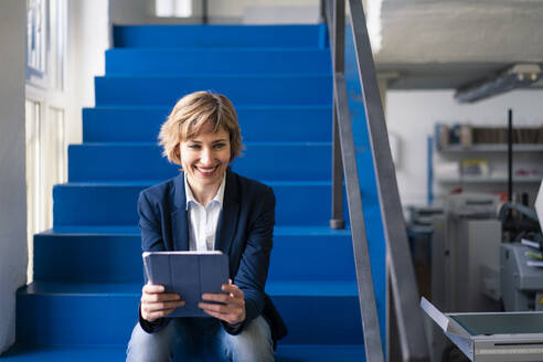 Lächelnde Geschäftsfrau, die ein digitales Tablet benutzt, während sie auf einer Treppe sitzt - JOSEF04185