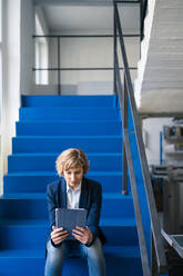Eine Unternehmerin benutzt ein digitales Tablet, während sie auf den Stufen einer Fabrik sitzt - JOSEF04187