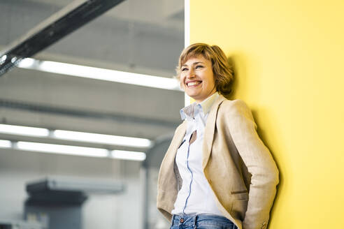 Lächelnde Geschäftsfrau, die sich an eine gelbe Wand in einer Druckerei lehnt - JOSEF04217
