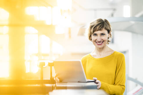 Lächelnde Geschäftsfrau mit Laptop in einer Druckwerkstatt - JOSEF04282