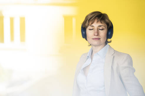Nachdenkliche Geschäftsfrau mit Kopfhörern vor einer gelben Wand - JOSEF04292