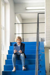 Lächelnde Besitzerin, die ein digitales Tablet benutzt, während sie auf den Stufen einer Werkstatt sitzt - JOSEF04304