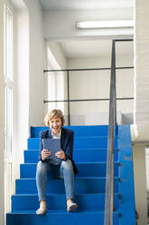 Smiling female owner using digital tablet while sitting on steps in workshop - JOSEF04304