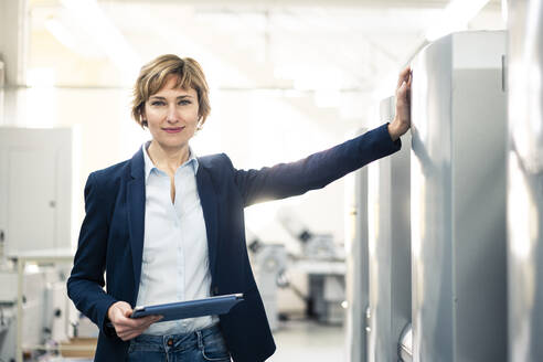 Smiling female manager standing with digital table by printing machine - JOSEF04310