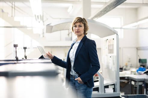 Businesswoman with hand in pocket standing by digital tablet in factory - JOSEF04318