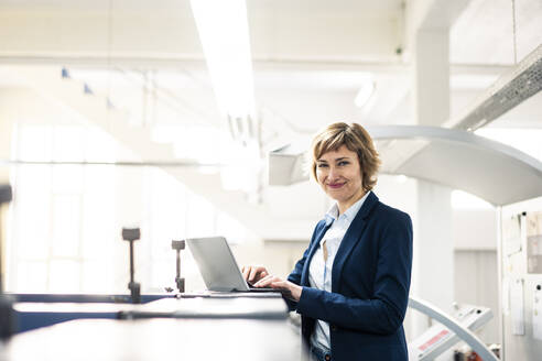 Smiling businesswoman standing by laptop in printing workshop - JOSEF04320