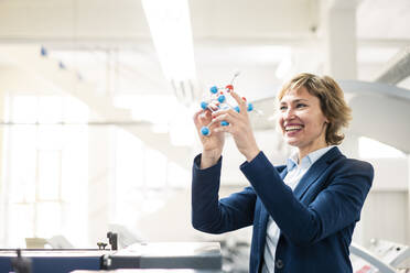 Lächelndes weibliches Fachwissen, das ein Molekularstrukturmodell hält, während es in einer Fabrik steht - JOSEF04325