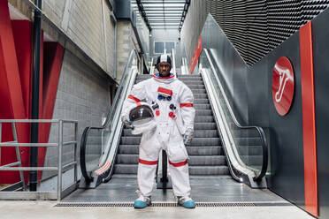 Mid adult man holding space helmet while standing in front of escalator - MEUF02690