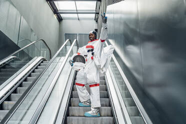 Fröhlicher Astronaut, der beim Besteigen einer Rolltreppe das Friedenszeichen macht - MEUF02694