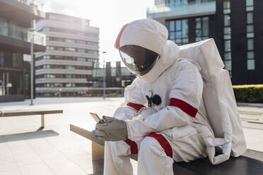 Männlicher Astronaut im Raumanzug, der ein Mobiltelefon benutzt, während er auf einer Bank sitzt - MEUF02705