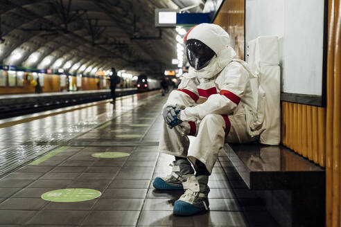 Männlicher Astronaut sitzt auf einer Bank am Bahnsteig - MEUF02763