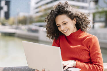 Lächelnde junge Frau, die an einem sonnigen Tag einen Laptop am Geländer benutzt - JCZF00697