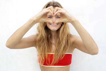 Smiling woman making heart shape gesture in front of white wall - OIPF00632