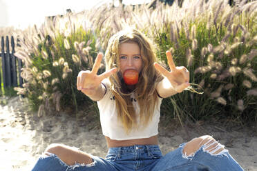 Woman eating apple showing peace sign while sitting on sand during sunny day - OIPF00638