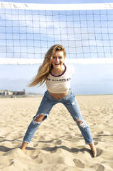 Playful young woman leaning on volleyball net at beach during sunny day - OIPF00647