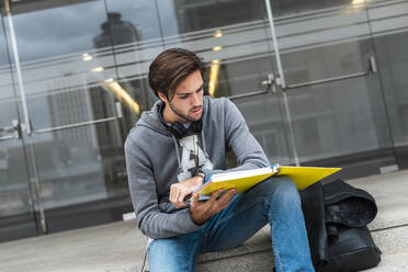 Junger Mann, der auf einer Treppe auf dem Campus sitzt und in einem Notizbuch liest - JMPF00952