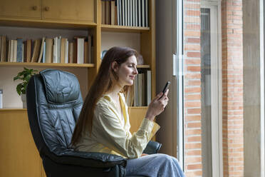 Lächelnde Frau, die ein Mobiltelefon auf einem Stuhl zu Hause benutzt - AFVF08734