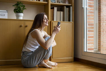 Young smiling female in shirt and panties using modern smartphone while  sitting on cozy bed in light bedroom stock photo