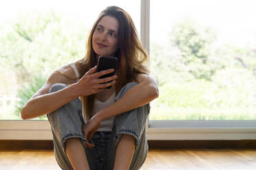 Smiling woman looking away while sitting in front of window - AFVF08741