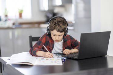 Boy wearing headphones writing on book while studying at home - WPEF04365