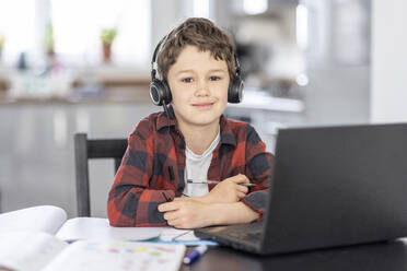 Lächelnder Junge mit Kopfhörern, der zu Hause vor einem Laptop sitzt - WPEF04366