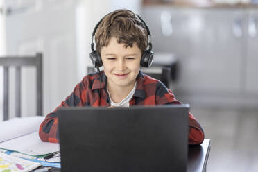 Smiling boy attending online classes through laptop while e-learning at home - WPEF04371