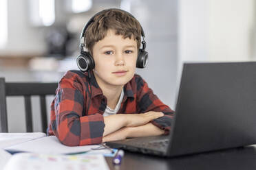 Boy wearing headphones e-learning while sitting with laptop at home - WPEF04373