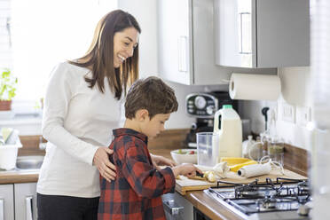 Lächelnde Mutter hilft ihrem Sohn beim Schneiden von Bananen in der Küche zu Hause - WPEF04396