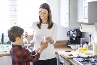 Lächelnde Mutter gibt ihrem Sohn in der Küche zu Hause ein Glas Milchshake - WPEF04402