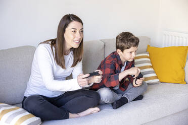 Curious mother playing video game by son on sofa at home - WPEF04411