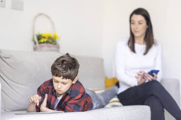 Junge, der ein digitales Tablet benutzt, während er auf dem Sofa liegt, mit seiner Mutter im Hintergrund zu Hause - WPEF04417