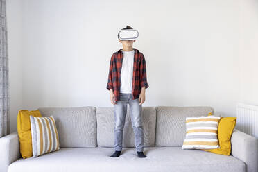 Boy wearing virtual reality headset standing on sofa in living room - WPEF04419