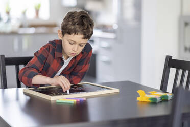 Boy drawing with chalks on a slate at table - WPEF04423