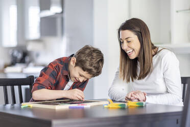 Lächelnde Mutter betrachtet ihren Sohn beim Zeichnen auf einer Schiefertafel in der Küche - WPEF04425