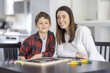 Smiling boy sitting by mother at table in kitchen - WPEF04426