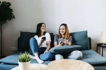 Lächelnde Freundinnen, die zu Hause auf dem Sofa sitzen und ein Mobiltelefon benutzen - XLGF01757